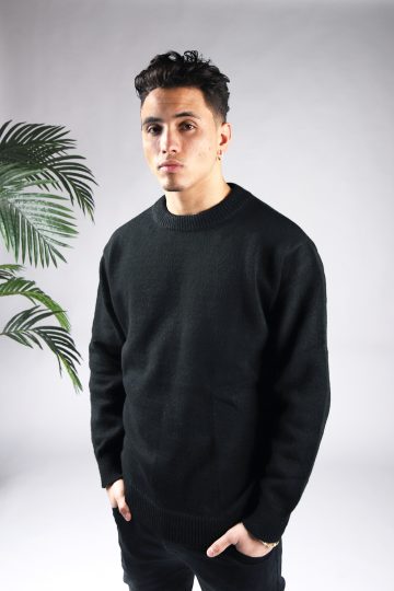 Vooraanzicht van model gekleed in een zwarte knit sweater in combinatie met een zwarte broek. Het model heeft zijn handen in zijn voorzakken en kijkt recht in de camera.