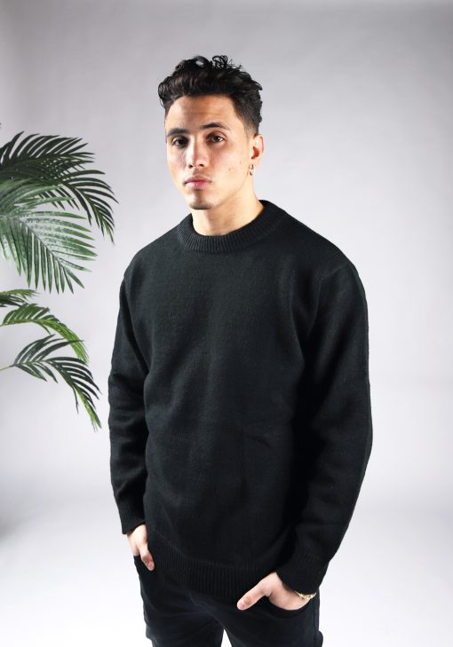 Vooraanzicht van model gekleed in een zwarte knit sweater in combinatie met een zwarte broek. Het model heeft zijn handen in zijn voorzakken en kijkt recht in de camera.