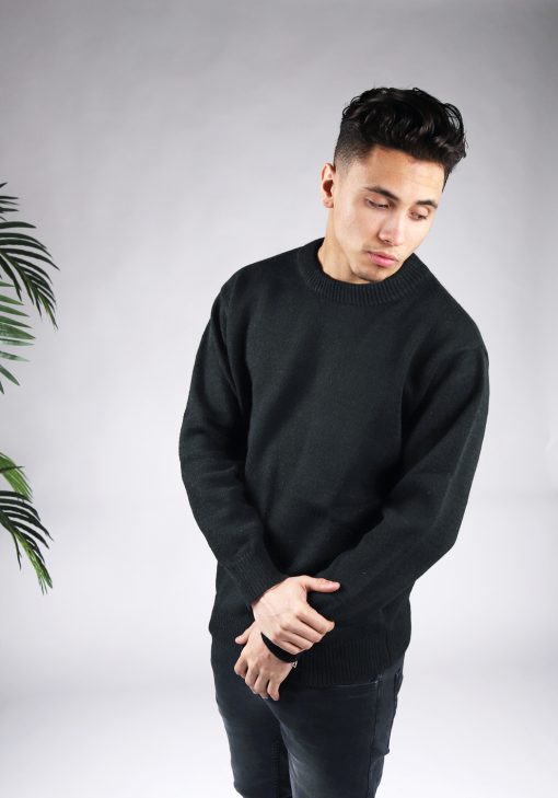 Vooraanzicht van model gekleed in een zwarte knit sweater in combinatie met een zwarte broek. Het model heeft zijn handen gekruist voor zich en kijkt schuin naar de grond.