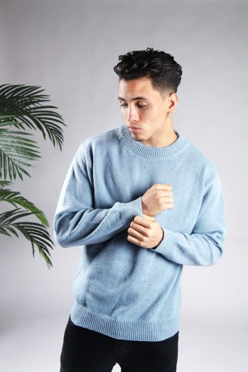 Vooraanzicht van model gekleed in een lichtblauwe knit sweater met een oversized fit in combinatie met een zwarte broek. Het model heeft zijn handen voor zijn borst en kijkt schuin naar de grond.