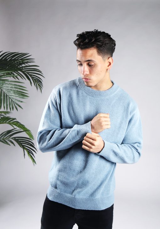 Vooraanzicht van model gekleed in een lichtblauwe knit sweater met een oversized fit in combinatie met een zwarte broek. Het model heeft zijn handen voor zijn borst en kijkt schuin naar de grond.
