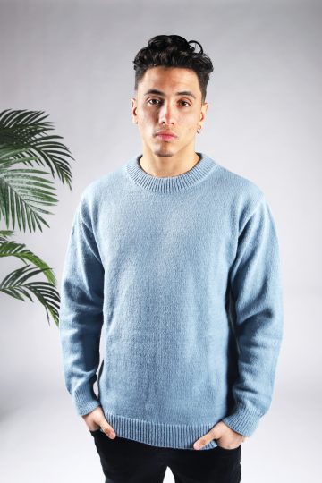 Vooraanzicht van model gekleed in een lichtblauwe knit sweater met een oversized fit in combinatie met een zwarte broek. Het model heeft zijn handen in zijn voorzakken en kijkt recht in de camera.