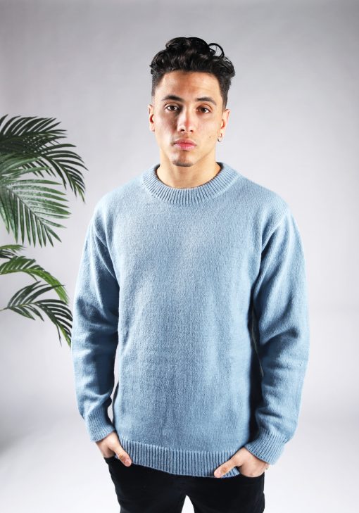 Vooraanzicht van model gekleed in een lichtblauwe knit sweater met een oversized fit in combinatie met een zwarte broek. Het model heeft zijn handen in zijn voorzakken en kijkt recht in de camera.