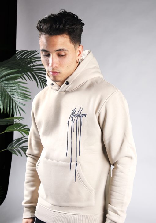 Vooraanzicht van model gekleed in beige hoodie met het donkerblauwe Malelions drip logo op de linkerborst. Het model heeft zijn armen langs zich en kijkt schuin naar de grond.