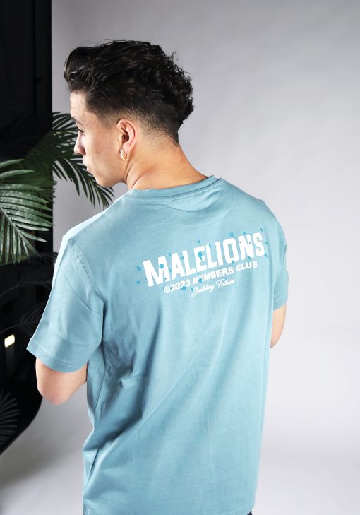 Schuin achteraanzicht van model gekleed in blauw Malelions t-shirt met witte tekst en blauwe verfspetters op de rug en linkerborst.