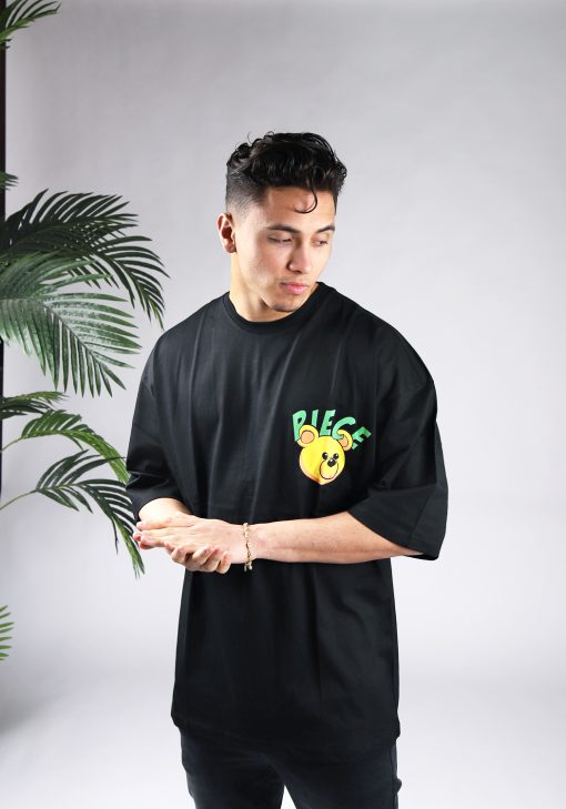 Vooraanzicht van een heren T-shirt in zwarte kleur en een super oversized pasvorm. Het T-shirt is voorzien van groene tekst en het hoofd van een beer op de linkerborst. Het model wrijft in zijn handen, kijkt naar linksonder en draagt een donkere jeans.