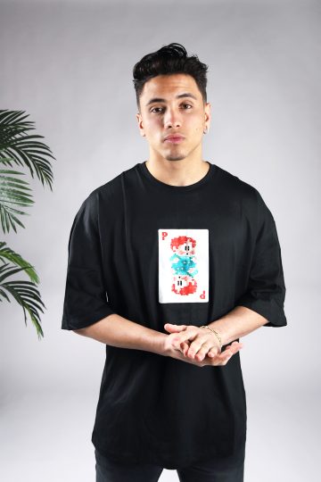 Vooraanzicht van een heren T-shirt in zwarte kleur en een super oversized pasvorm. Het T-shirt is voorzien van een pixelart speelkaart met daarop een pixelart karakter op de voorkant. Het model heeft wrijft in zijn handen en draagt een donkere jeans.