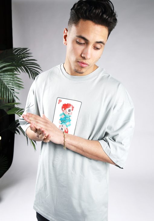 Vooraanzicht van een heren T-shirt in grijze kleur en een super oversized pasvorm. Het T-shirt is voorzien van een pixelart speelkaart met daarop een pixelart karakter op de voorkant. Het model wrijft in zijn handen en kijkt naar linksonder.