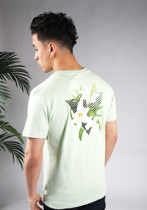 Schuin achteraanzicht van model gekleed in lichtgroen t-shirt met driekhoek bloemenprint op de rug, en het kleine zwarte logo op de borst.