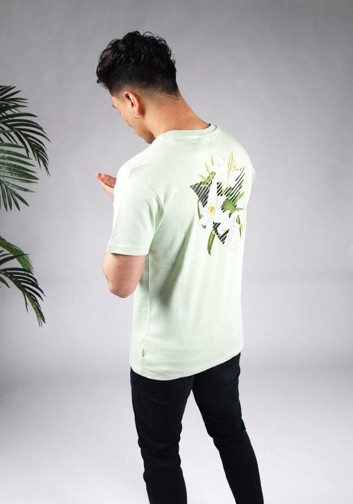 Schuin achteraanzicht van model gekleed in lichtgroen t-shirt met driekhoek bloemenprint op de rug, en het kleine zwarte logo op de borst.