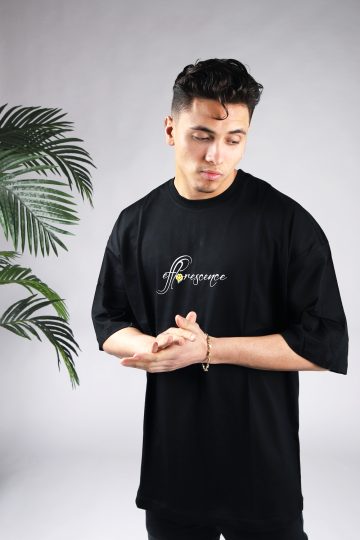 Vooraanzicht van een heren T-shirt in zwarte kleur in een oversized pasvorm. Het T-shirt is voorzien van tekst op de borst. Het model wrijft in zijn handen en kijkt naar linksonder.