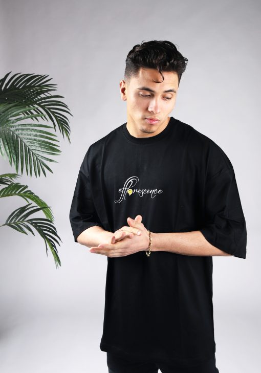 Vooraanzicht van een heren T-shirt in zwarte kleur in een oversized pasvorm. Het T-shirt is voorzien van tekst op de borst. Het model wrijft in zijn handen en kijkt naar linksonder.
