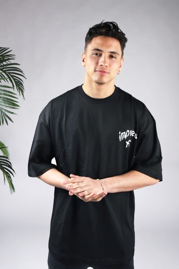 Vooraanzicht van een heren T-shirt in zwarte kleur en een super oversized pasvorm. Het T-shirt is voorzien van witte tekst op de linkerborst. Het model wrijft in zijn handen.