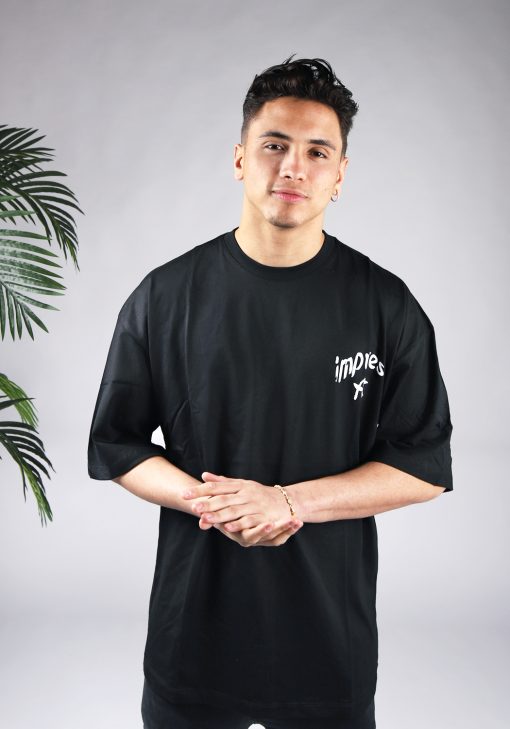 Vooraanzicht van een heren T-shirt in zwarte kleur en een super oversized pasvorm. Het T-shirt is voorzien van witte tekst op de linkerborst. Het model wrijft in zijn handen.