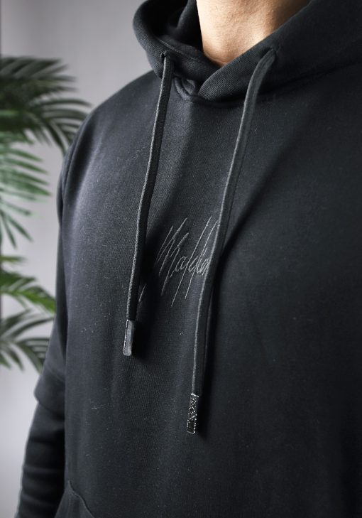 Close up van model gekleed in zwarte hoodie met het zwarte geborduurde Malelions logo in het midden van de borst en zwarte hoodie strings.