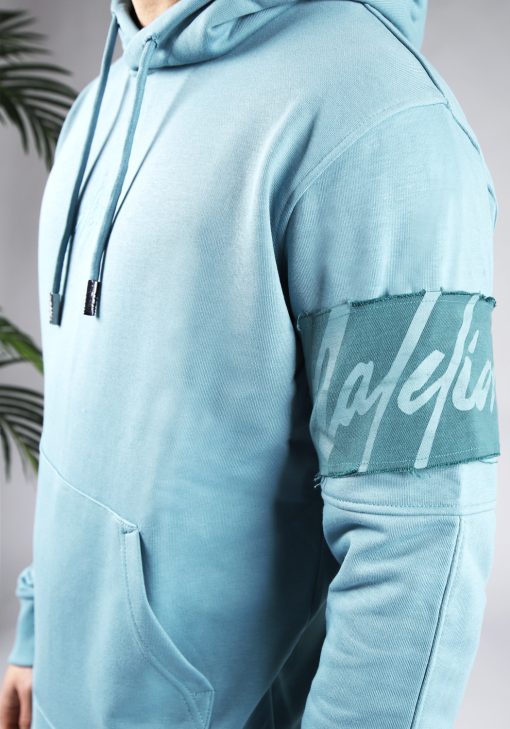 Close up van blauwe hoodie met het blauwe geborduurde Malelions logo in het midden van de borst. De trui heeft een brede turquoise band om de linker bovenarm waarop het lichtblauw logo staat.