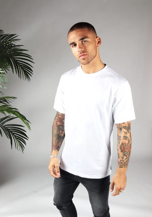 Linker vooraanzicht van heren T-shirt in witte kleur, met ronde hals en oversized pasvorm. Het model draagt een donkere jeans en heeft zijn armen langs zijn lichaam.