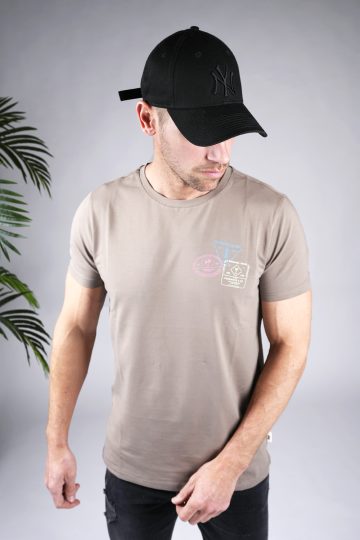 Vooraanzicht van model gekleed in zandkleurig shirt met lichtgele surf print op de rug en gekleurde print op de linkerborst. Het model draagt een zwarte broek en een zwarte pet.