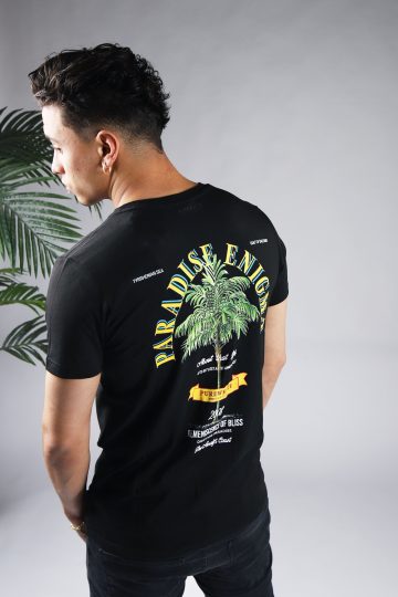 Schuin achteraanzicht van model gekleed in zwarte paradise tee met een print van een palmboom omringd met tekst op de rug en een klein logo op de voorkant.