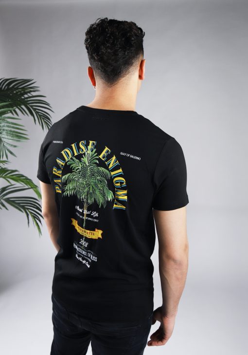 Achteraanzicht van model gekleed in zwarte paradise tee met een print van een palmboom omringd met tekst op de rug en een klein logo op de voorkant.