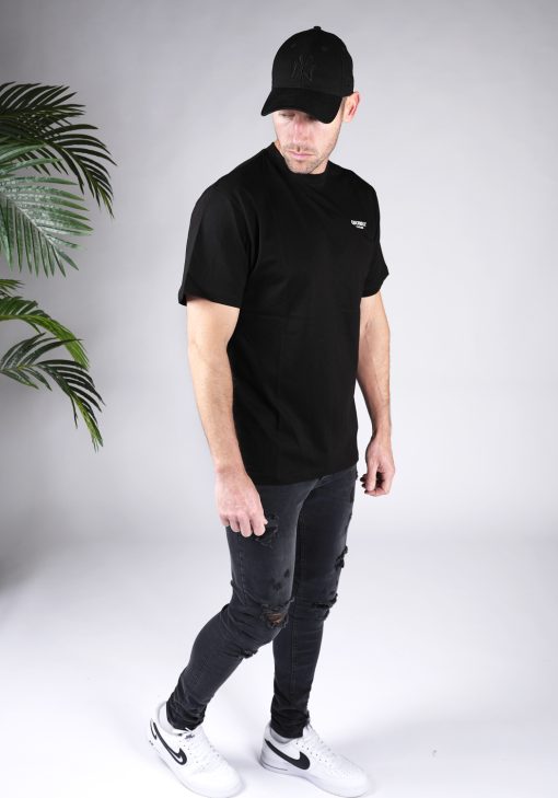 Vooraanzicht van heren T-shirt in zwarte kleur, met ronde hals en een relaxed fit pasvorm. Het T-shirt is voorzien van het QUOTRELL-logo in witte kleur op de borst. Het model combineert het T-shirt met witte sneakers.