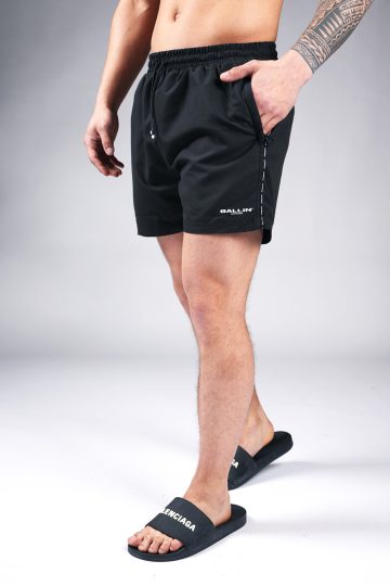 Zijaanzicht van model gekleed in zwarte Ballin swimshorts met het witte logo op de linkerbeen. Het model heeft een hand in zijn zak en draagt zwarte badslippers.