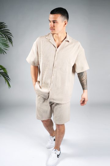 Vooraanzicht van model gekleed in beige blouse set met lichte rib stof. De set bestaat uit shorts en een blouse met korte mouwen. Het model heeft een hand in zijn zak en kijkt schuin naar rechts.