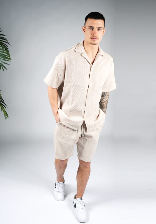 Vooraanzicht van model gekleed in beige blouse set met lichte rib stof. De set bestaat uit shorts en een blouse met korte mouwen. Het model heeft zijn handen achter zich en kijkt recht in de camera.