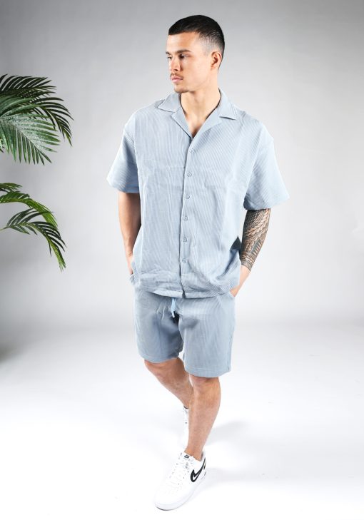 Vooraanzicht van model gekleed in lichtblauwe blouse set met lichte rib stof. De set bestaat uit shorts en een blouse met korte mouwen. Het model heeft zijn handen in zijn zakken en kijjkt naar rechts.