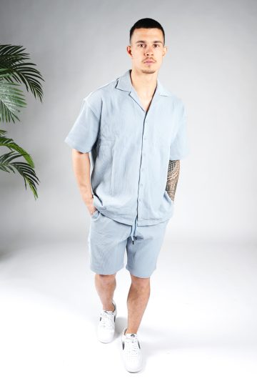 Vooraanzicht van model gekleed in lichtblauwe blouse set met lichte rib stof. De set bestaat uit shorts en een blouse met korte mouwen. Het model heeft zijn handen in zijn zakken en kijkt recht in de camera.