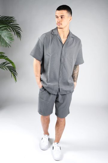 Vooraanzicht van model gekleed in grijze blouse set met lichte rib stof. De set bestaat uit shorts en een blouse met korte mouwen. Het model heeft zijn handen in zijn zakken en kijkt naar rechts.