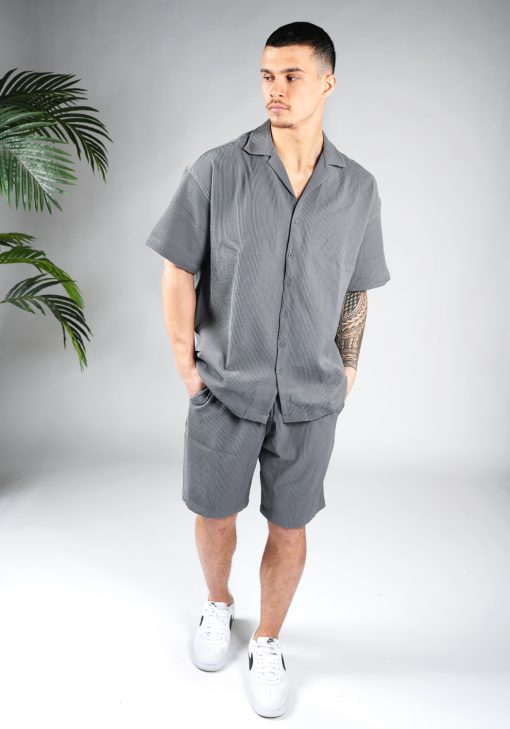 Vooraanzicht van model gekleed in grijze blouse set met lichte rib stof. De set bestaat uit shorts en een blouse met korte mouwen. Het model heeft zijn handen in zijn zakken en kijkt naar rechts.