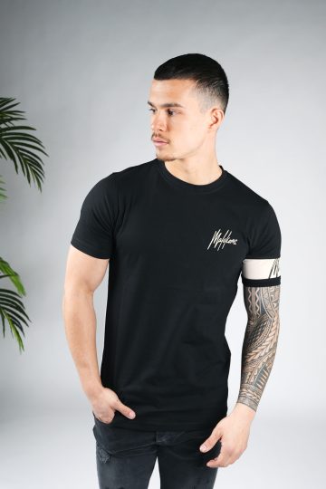 Vooraanzicht van model gekleed in zwart t-shirt met het zandkleurige logo op de linkerborst en een zandkleurige band om de linkerarm met daarop het zwarte logo. Het model heeft een hand in zijn zak en kijkt naar rechts.