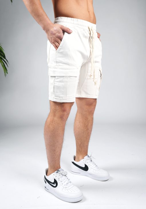 Rechter zijaanzicht van model gekleed in zandkleurige cargo shorts in combinatie met witte sneakers. Het model heeft zijn handen in zijn zakken.