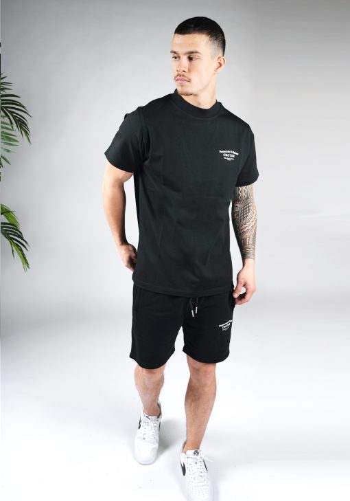 Vooraanzicht van model gekleed in zwarte CROYEZ set. De set bestaat uit jogger shorts met witte tekst op de linkerbovenbeen en een t-shirt met tekst op de rug en linkerborst.