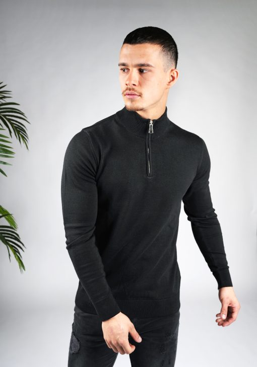 Model met armen langs zijn lichaam draagt een zwarte trui met halve rits gecombineerd met een zwarte broek. Model kijkt schuin naar rechts.