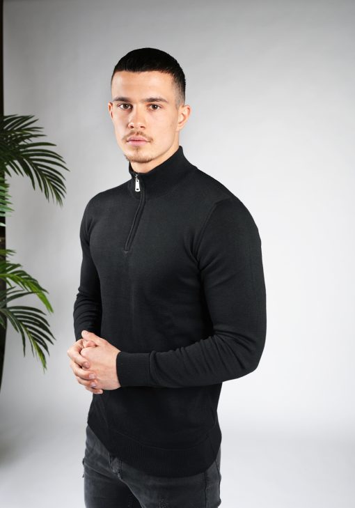 Model met handen over elkaar draagt een zwarte trui met halve rits gecombineerd met een zwarte broek.