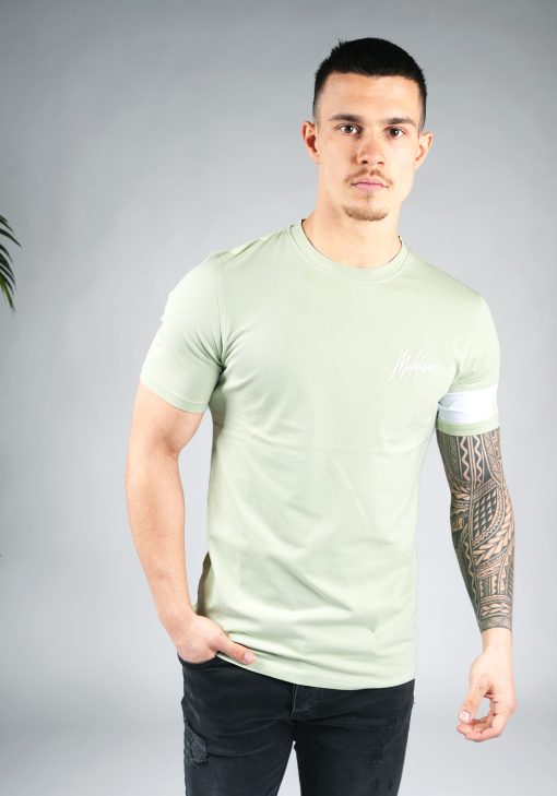 Vooraanzicht van model gekleed in lichtgroen t-shirt met het witte logo op de linkerborst en een witte band om de linkerarm met daarop het groene logo. Het model heeft een arm in zijn zak en kijkt recht in de camera.