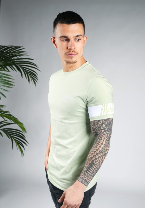 Zijaanzicht van model gekleed in lichtgroen t-shirt met het witte logo op de linkerborst en een witte band om de linkerarm met daarop het groene logo. Het model heeft een arm in zijn zak en kijkt schuin naar de grond.