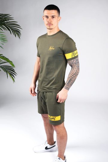 Schuin vooraanzicht van model gekleed in legergroene Malelions set met gele details. De set bestaat uit een shirt met het gele Malelions logo op de borst en een gele band om de linker arm, en de jogger shorts met de gele band om de linkerbeen.