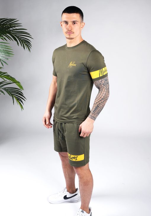 Schuin vooraanzicht van model gekleed in legergroene Malelions set met gele details. De set bestaat uit een shirt met het gele Malelions logo op de borst en een gele band om de linker arm, en de jogger shorts met de gele band om de linkerbeen.