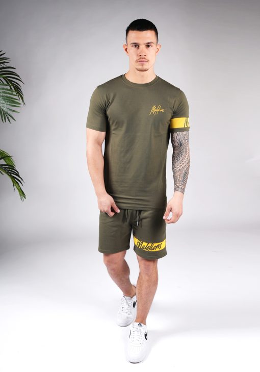 Vooraanzicht van model gekleed in legergroene Malelions set met gele details. De set bestaat uit een shirt met het gele Malelions logo op de borst en een gele band om de linker arm, en de jogger shorts met de gele band om de linkerbeen.
