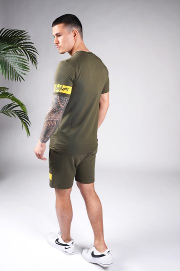 Achteraanzicht van model gekleed in legergroene Malelions set met gele details. De set bestaat uit een shirt met het gele Malelions logo op de borst en een gele band om de linker arm, en de jogger shorts met de gele band om de linkerbeen.