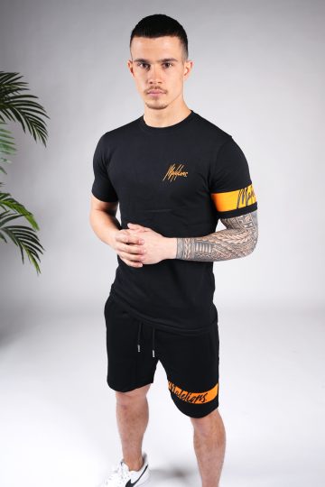 Vooraanzicht van model gekleed in zwarte Malelions set met oranje details. De set bestaat uit een shirt met het oranje Malelions logo op de borst en een oranje band om de linker arm, en de jogger shorts met de oranje band om de linkerbeen. Het model heeft zijn handen voor zich.