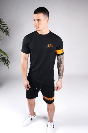 Vooraanzicht van model gekleed in zwarte Malelions set met oranje details. De set bestaat uit een shirt met het oranje Malelions logo op de borst en een oranje band om de linker arm, en de jogger shorts met de oranje band om de linkerbeen.
