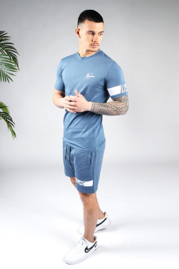Schuin vooraanzicht van model gekleed in blauwe Malelions set met witte details. De set bestaat uit een shirt met het witte Malelions logo op de borst en een witte band om de linker arm, en de jogger shorts met de witte band om de linkerbeen.