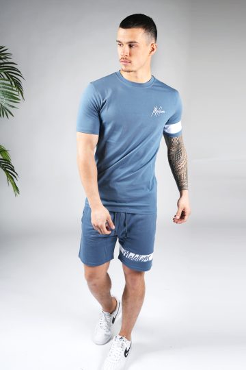 Vooraanzicht van model gekleed in blauwe Malelions set met witte details. De set bestaat uit een shirt met het witte Malelions logo op de borst en een witte band om de linker arm, en de jogger shorts met de witte band om de linkerbeen.