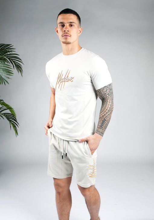 Schuin vooraanzicht van model gekleed in lichtgrijze Malelions set. De set bestaat uit een shirt met het dubbelkleurige geborduurde Malelions logo in het grijs en oranje, en de shorts met hetzelfde logo op de been.
