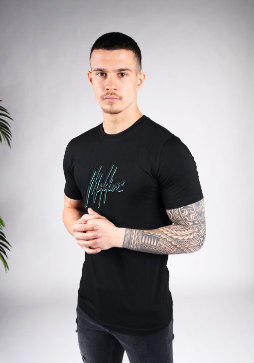 Schuin vooraanzicht van model gekleed in zwart Malelions t-shirt met het dubbel geborduurde logo in de kleuren turquoise en zwart op de borst. Het model heeft zijn handen voor zich en kijkt recht in de camera.