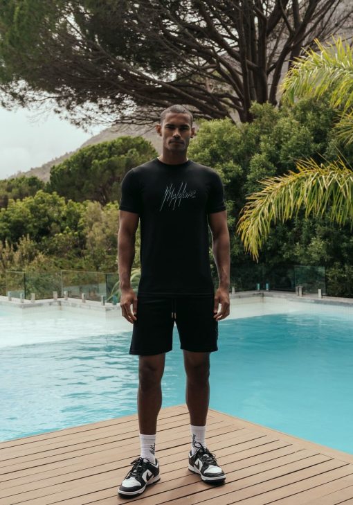 Vooraanzicht van model gekleed in zwart Malelions t-shirt met het dubbel geborduurde logo in de kleuren turquoise en zwart op de borst. Het model staat op een tropische locatie langs het zwembad.